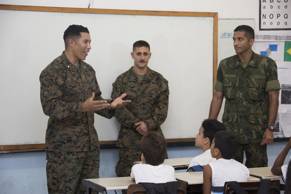 Three military personnel in camouflage uniforms are visiting a classroom and addressing a group of children seated at desks. Two individuals appear to be wearing US Marine Corps MARPAT digital camouflage uniforms, while the third wears what appears to be a Brazilian military uniform with a Brazilian flag patch visible on the wall behind them. The leftmost soldier, whose name tape reads 'REAMON,' is actively speaking and gesturing to the students.