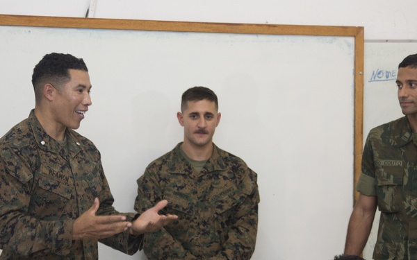 Three military personnel in camouflage uniforms are visiting a classroom and addressing a group of children seated at desks. Two individuals appear to be wearing US Marine Corps MARPAT digital camouflage uniforms, while the third wears what appears to be a Brazilian military uniform with a Brazilian flag patch visible on the wall behind them. The leftmost soldier, whose name tape reads 'REAMON,' is actively speaking and gesturing to the students.
