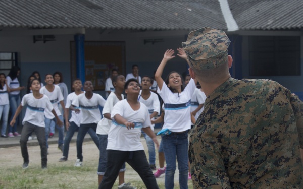 A military service member in digital camouflage uniform interacts with a large group of children in what appears to be a school courtyard. The children, wearing matching white t-shirts with blue stripes, appear joyful and energetic, some with arms raised. The setting suggests a community outreach or humanitarian assistance event at a school facility.