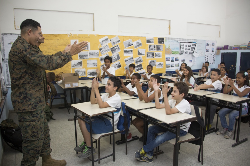 A military service member in camouflage uniform is engaging with a classroom full of elementary school-aged children who are clapping and raising their hands enthusiastically. The setting appears to be a school classroom with student project displays mounted on the back wall. The interaction appears positive and energetic, suggesting a community outreach or educational visit.