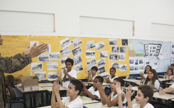 A military service member in camouflage uniform is engaging with a classroom full of elementary school-aged children who are clapping and raising their hands enthusiastically. The setting appears to be a school classroom with student project displays mounted on the back wall. The interaction appears positive and energetic, suggesting a community outreach or educational visit.