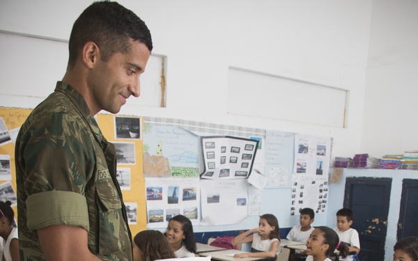 A soldier in Brazilian military camouflage uniform is distributing notebooks or booklets to students in a classroom setting. A young girl in the foreground is smiling as she receives a booklet featuring a globe logo. The classroom appears to be in Brazil, with multiple children seated at individual desks.