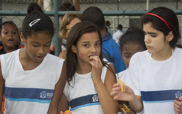 Three girls wearing matching white uniforms with 'Rio Prefeitura Educação' (Rio de Janeiro municipal education) branding are gathered around a table with plates of baked goods and pastries. The middle girl is eating a snack while holding a cupcake, and the others are selecting items from the spread. The setting appears to be a school event or food distribution activity in what looks like an outdoor or semi-outdoor school facility.