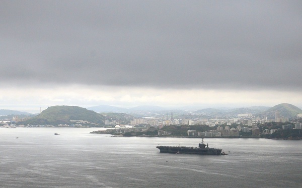 An elevated aerial view of a large bay or harbor, likely Guanabara Bay in Rio de Janeiro, Brazil, identifiable by the characteristic geography and urban skyline in the background. A large aircraft carrier is visible navigating the bay, accompanied by an offshore oil drilling platform anchored to the left. The scene is captured under overcast, cloudy conditions giving the image a grey, muted tone.