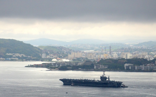 A large aircraft carrier is navigating through a bay or harbor, accompanied by smaller tugboats, with a coastal city and green hills visible in the background. The scene appears to be taken from an elevated vantage point, possibly a hilltop or tall structure. Overcast and hazy weather conditions reduce visibility in the distance.