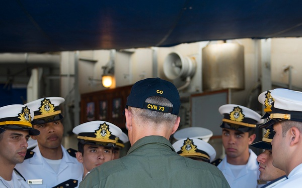 A U.S. Navy Commanding Officer (CO) of CVN-73, wearing an olive green flight suit and a black cap labeled 'CO CVN 73', addresses a group of foreign naval officers in white dress uniforms. The foreign officers appear to be from a Latin American navy, identifiable by their distinctive cap insignia and uniform style. The setting appears to be aboard a U.S. Navy aircraft carrier, likely the USS George Washington (CVN-73).