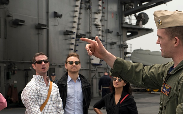 A U.S. Navy or Marine Corps officer in a green flight suit is conducting a tour on what appears to be the flight deck of an aircraft carrier. He is pointing and explaining something to a group of four civilian visitors, one of whom is photographing with a smartphone. The setting is an active or docked naval vessel with large ship structures visible in the background.
