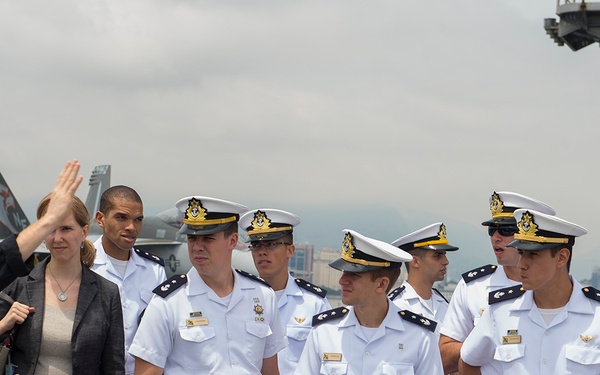 A group of naval officers in white dress uniforms with gold-emblem caps are gathered on what appears to be the flight deck of an aircraft carrier. A person in dark civilian clothing is gesturing and appears to be briefing or guiding the group, accompanied by a woman in business attire. Military aircraft are visible in the background, along with an overcast sky and a waterfront cityscape.