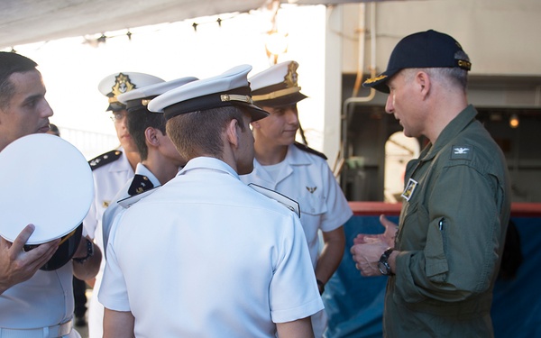 A group of naval personnel in white dress uniforms are engaged in conversation with a person wearing an olive/green flight suit and dark cap. The setting appears to be on or near a naval vessel, indicated by the ship structure visible in the background. One officer is holding a white naval cap while speaking with the flight-suited individual.