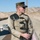A U.S. Marine in MARPAT digital camouflage uniform stands at what appears to be a rifle range in a desert environment. He is wearing a tan shooting jacket with padded shoulders and multiple buckle closures, and is holding an M16/M4-style rifle. Numbered target markers are visible in the background, confirming a marksmanship training setting.
