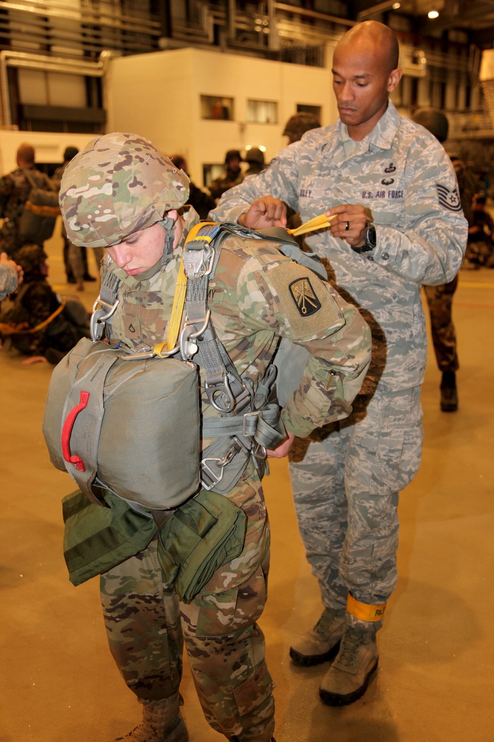 A U.S. Air Force Technical Sergeant is conducting a pre-jump equipment inspection on a U.S. Army soldier wearing a full parachute rig and combat gear. The scene takes place inside what appears to be a military hangar or staging area, with additional personnel visible in the background. This appears to be a standard jumpmaster personnel inspection (JMPI) prior to an airborne operation.
