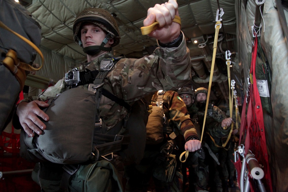 Military paratroopers are lined up inside a military transport aircraft, preparing for a jump operation. The lead soldier in the foreground is holding a static line snap hook, indicating imminent exit from the aircraft. Multiple soldiers wearing different camouflage patterns are visible, suggesting a multinational or joint military exercise.