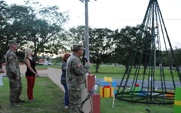 A military ceremony or event is taking place outdoors on a grassy area, likely a Christmas tree lighting ceremony on a military installation. A soldier in camouflage uniform is speaking into a microphone at a podium, while a small group of military personnel and civilians stand nearby. Large decorative gift box props and what appears to be a wire-frame Christmas tree structure are visible in the background.