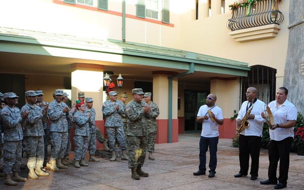 A group of military personnel in camouflage uniforms are gathered in an outdoor courtyard, applauding a small civilian musical ensemble. The musicians are dressed in white shirts and are playing instruments including saxophones and a tambourine/pandeiro. The setting appears festive, with Christmas decorations visible including poinsettias and holiday wreaths, suggesting a holiday celebration event.