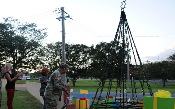 A military Christmas tree lighting ceremony is taking place outdoors on a grassy area, likely on a military installation. Two soldiers in camouflage uniforms are present alongside several civilians, with one soldier appearing to activate the lights on a large wire-frame Christmas tree. Decorative oversized gift boxes are arranged around the base of the tree at dusk.