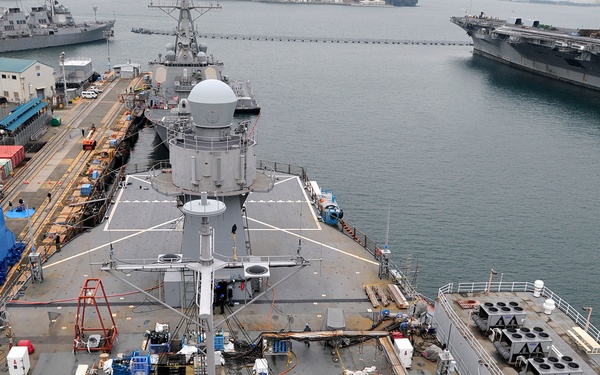 An aerial view of a naval vessel undergoing maintenance or refit operations at a shipyard, likely a US Navy destroyer or cruiser. The ship's deck is covered with equipment, hoses, cables, and personnel performing various maintenance tasks. In the background, additional naval vessels including what appears to be an aircraft carrier are visible in the harbor.