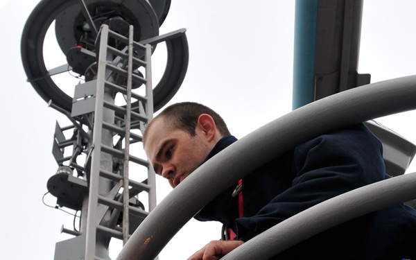 Two workers in dark uniforms are performing maintenance or inspection work at elevation on what appears to be a naval vessel's mast or communications tower. Both individuals are wearing safety harnesses with blue straps, indicating adherence to fall protection protocols. The photo is taken from a low angle looking upward, emphasizing the height at which they are working.