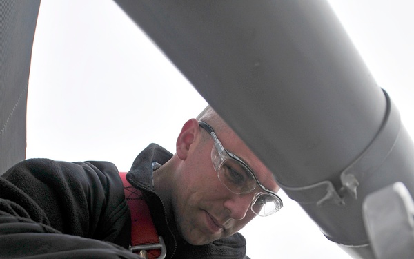 A technician wearing a red safety harness and clear protective eyeglasses is performing maintenance or inspection work on what appears to be a large piece of industrial or aerospace equipment. The low-angle photograph captures the worker leaning over a mechanical component, using his hands to manipulate a fastener or fitting. The overcast sky is visible in the background, suggesting the work is being performed outdoors at elevation.