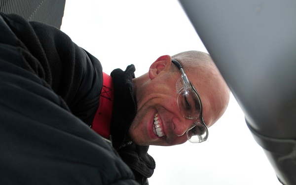 A bald man wearing safety glasses and a black jacket with a red safety harness is performing maintenance or technical work on mechanical equipment at height. He is smiling broadly while using his hands to manipulate a component on what appears to be aircraft or elevated industrial equipment. A yellow safety lanyard is visible, indicating proper fall protection protocols are in use.