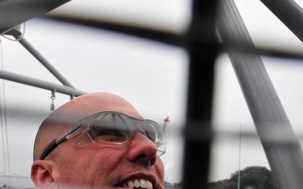 A bald man wearing clear safety glasses is photographed looking upward through a metal scaffolding or structural framework. The image is taken from a low angle through the grid of metal bars, creating a framed, layered composition. A waterfront or harbor environment is visible in the background under an overcast sky.