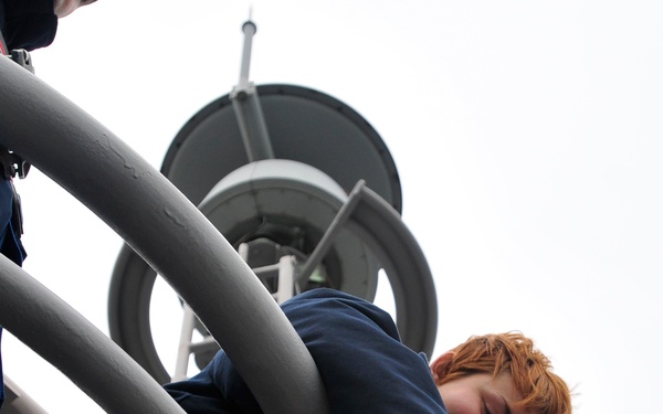 A young person with red/auburn hair wearing a navy blue shirt is positioned at height on what appears to be a ship's mast or communications tower structure. They are equipped with a safety harness in red and blue, secured with multiple buckles and straps. The photo is taken from a low angle looking upward, with circular radar or communications equipment visible above.