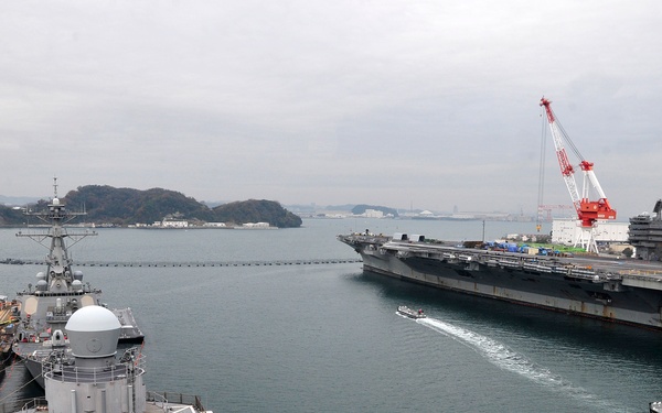 A naval base harbor scene showing multiple warships docked and maneuvering in what appears to be Yokosuka Naval Base, Japan. A large aircraft carrier bearing the hull number '76' (USS Ronald Reagan, CVN-76) is prominently visible on the right side, alongside smaller destroyers and support vessels. Industrial cranes painted red and white are visible in the background, along with forested hills characteristic of the Japanese coastline.