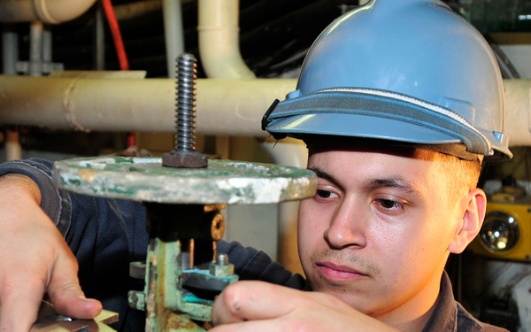A young male worker wearing a blue hard hat and dark coveralls is performing maintenance on a large industrial valve or pipe fitting in what appears to be a mechanical room or engine room. He is using a wrench to tighten or loosen a component on the heavily corroded, green-patinated valve assembly. The setting suggests a ship's engine room, industrial facility, or similar heavy-duty mechanical environment.