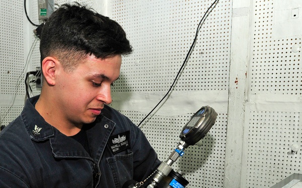 A U.S. Navy sailor in dark blue coveralls is examining or assembling a mechanical/optical instrument in what appears to be a shipboard workshop or maintenance space. The sailor's uniform displays a name tag and what appears to be a warfare qualification badge on the chest. The workspace includes a pegboard wall, filing cabinet, and various tools and equipment in the background.