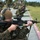 Military personnel are conducting a live-fire rifle range exercise at an outdoor shooting range. The foreground shows a Marine in MARPAT camouflage uniform aiming an M4 carbine with an ACOG optic while wearing hearing protection. Multiple additional shooters are visible in the background in kneeling positions along the firing line.