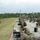 Military personnel, appearing to be U.S. Marines in woodland MARPAT camouflage uniforms, are conducting a live-fire rifle range qualification exercise at an outdoor shooting range. Multiple shooters are positioned in prone and kneeling stances along marked firing lanes, with range safety officers and observers standing behind them. The range appears to be located in a subtropical environment, consistent with installations in Okinawa, Japan.