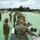 Military personnel in camouflage uniforms are conducting a pistol qualification or training exercise at an outdoor shooting range. Multiple shooters are lined up at firing positions along a long green-surfaced range, aiming downrange at targets. Support personnel and range safety officers are observing from behind the firing line.