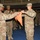 A military ceremony is taking place inside what appears to be a gymnasium or indoor arena, where two soldiers in MultiCam/OCP camouflage uniforms are handling an orange guidon or unit flag. Multiple soldiers stand in formation in the background, suggesting this is a formal unit ceremony such as a casing or uncasing of colors, activation, or deactivation event. Civilian spectators are visible in bleachers on the right side of the image.