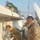Two military personnel in different camouflage uniforms are working on a rooftop installation, appearing to drill or route cables through a wall. One soldier wearing what appears to be US Army MultiCam uniform is actively using a power drill, while the other in a Spanish military pixelated camouflage pattern observes. The rooftop setting includes sandbags, satellite dishes, HVAC units, and multiple cables.