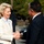 German, Turkish leadership meet to discuss coalition forces partnerships Two individuals are engaged in a formal handshake outdoors, likely at an official or diplomatic meeting. A woman in a white blazer with a light blue collar is smiling broadly at a man in a dark suit wearing glasses. A third person in a dark jacket is visible in the background.