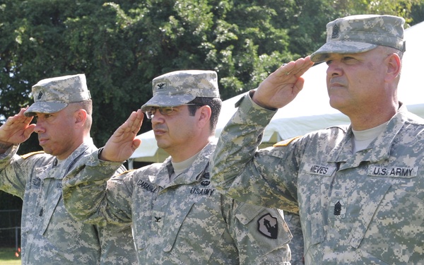 Three U.S. Army soldiers in ACU (Army Combat Uniform) digital camouflage are standing at attention and rendering a salute during what appears to be an outdoor military ceremony. The soldiers are aligned in a row on a grass field, with a white tent canopy visible in the background. Name tapes and unit patches are visible on their uniforms.