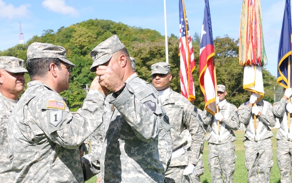 A U.S. Army military ceremony is taking place on a grass field, likely a change of command or award ceremony. A soldier in the foreground is having his cover (cap) adjusted or a pin placed on it by another soldier, while a color guard stands at attention in the background holding multiple flags including the U.S. flag and what appears to be a Texas state flag. Multiple soldiers in ACU (Army Combat Uniform) digital camouflage are present in formation.