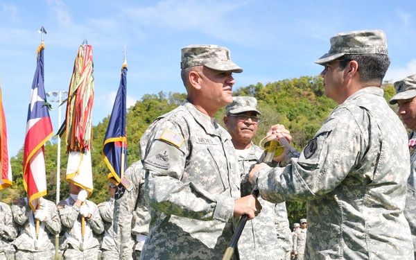 A formal U.S. Army military ceremony is taking place on a grass parade field. Two senior soldiers in the foreground appear to be exchanging or passing a guidon or ceremonial saber, consistent with a change of command ceremony. Troops in ACU (Army Combat Uniform) are standing in formation in the background alongside multiple flags including the U.S. flag and what appears to be a Puerto Rican flag.