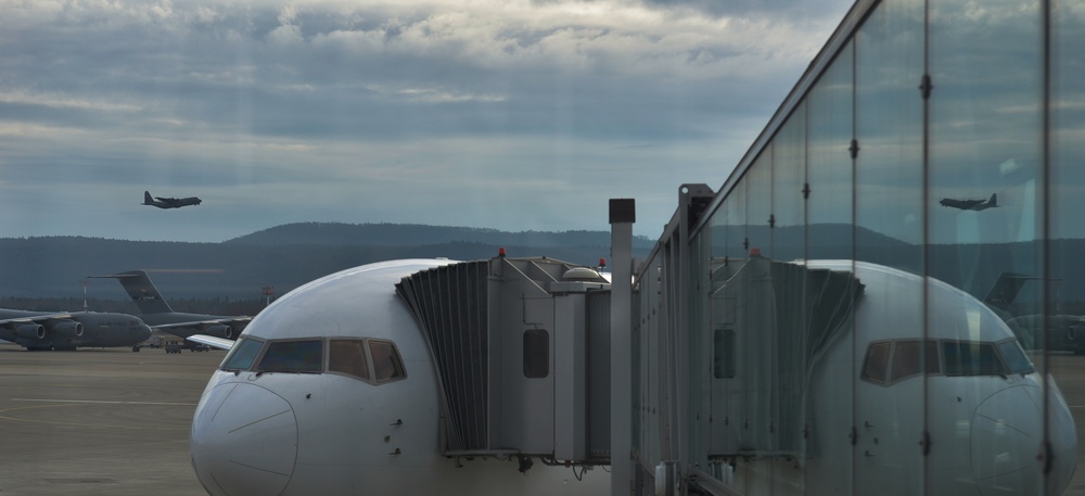 The image shows a commercial aircraft docked at a jet bridge at an airport terminal, photographed through a glass window that creates a mirror reflection of the scene. In the background, military transport aircraft are visible on the tarmac and in flight, suggesting this airport serves both civilian and military operations. The overcast sky and distant hills provide a moody atmospheric backdrop.