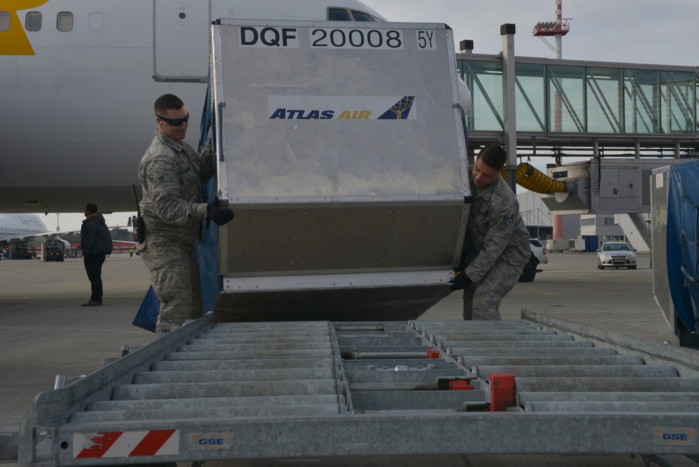 Two U.S. Air Force personnel in ABU (Airman Battle Uniform) camouflage are handling an Atlas Air cargo unit container (ULD) marked 'DQF 20008 5Y' on an airport tarmac. They are maneuvering the container onto or off of a ground support equipment (GSE) roller conveyor ramp adjacent to a commercial aircraft. The setting appears to be an international airport with multiple aircraft and terminal infrastructure visible in the background.