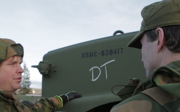 Two soldiers in Norwegian Army camouflage uniforms are interacting near a military vehicle marked 'USMC-638417' with 'DT' written in white chalk or paint. The soldier on the left is pointing at the vehicle while appearing to explain or brief the other soldier. The setting appears to be a cold, wintry outdoor environment, likely in Norway.