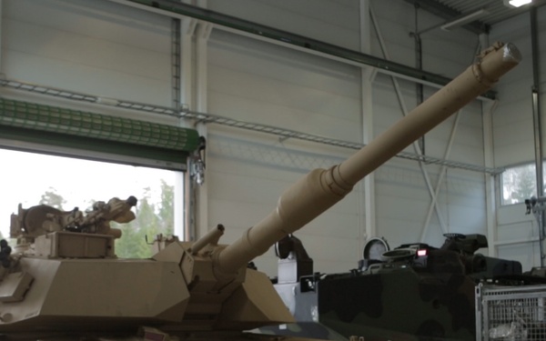 A tan/desert-camouflage painted M1 Abrams main battle tank is positioned inside a large military maintenance or storage hangar. The tank's main gun is elevated and pointed toward the upper left. The facility appears to be a modern, well-lit structure with additional military equipment visible in the background.
