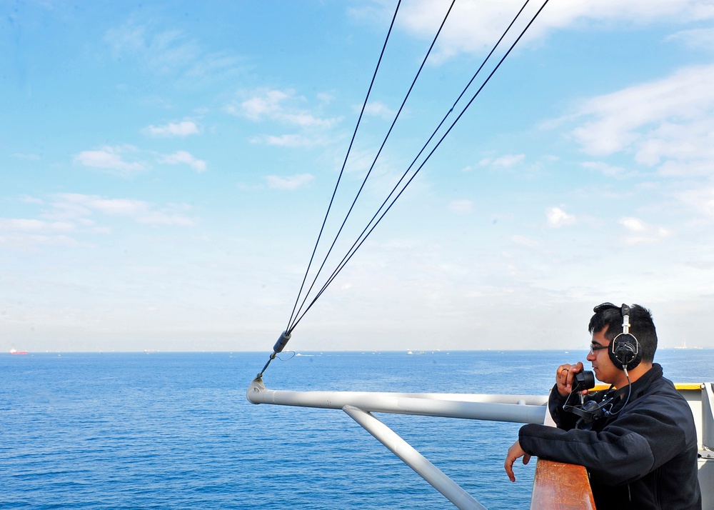 A person wearing headphones and sunglasses stands on the deck of a vessel, looking through binoculars toward the open sea. The scene shows calm blue ocean water under a partly cloudy sky, with distant ships visible on the horizon. The individual appears to be performing a lookout or observation duty, consistent with maritime operations.