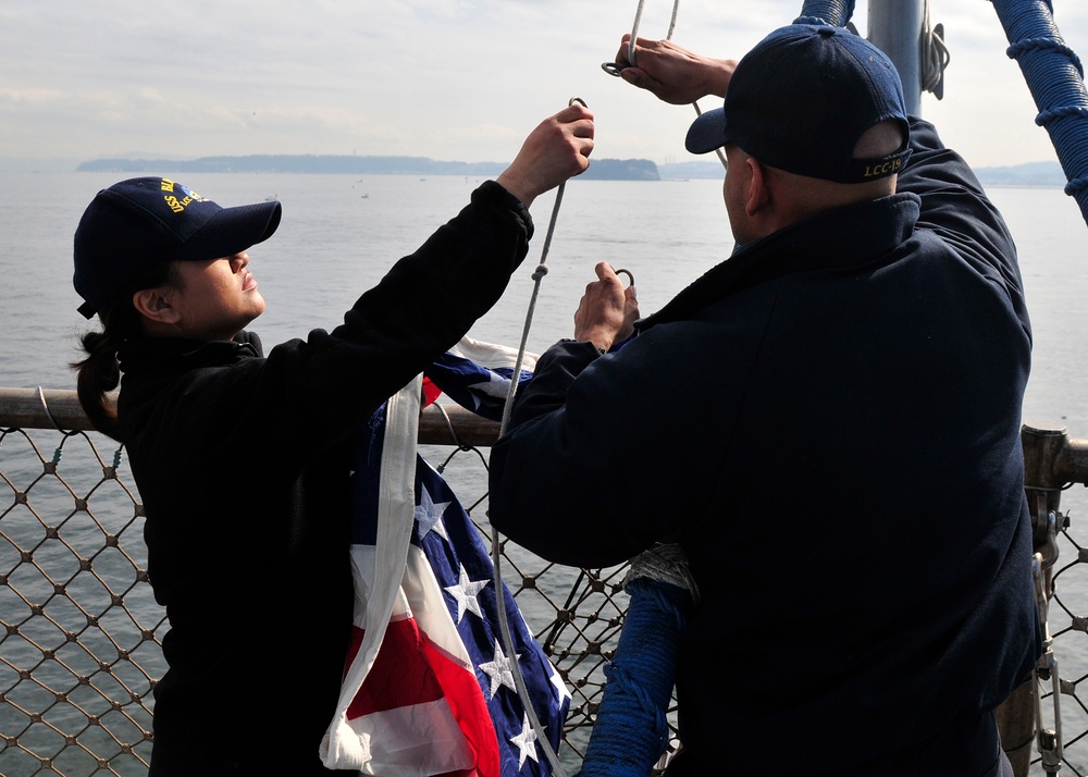 Two naval personnel in dark blue uniforms and caps are working together to attach or raise an American flag on a ship's mast or halyard. They are standing on the deck of a naval vessel, identifiable by the chain-link safety railing and rigging visible in the frame. The scene takes place on open water with a distant landmass visible on the horizon.