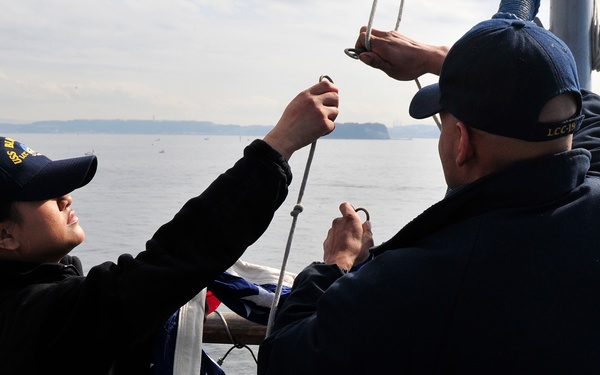 Two naval personnel in dark blue uniforms and caps are working together to attach or raise an American flag on a ship's mast or halyard. They are standing on the deck of a naval vessel, identifiable by the chain-link safety railing and rigging visible in the frame. The scene takes place on open water with a distant landmass visible on the horizon.