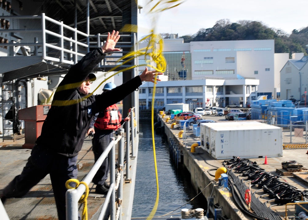 A sailor dressed in black is throwing a yellow heaving line from the deck of a naval vessel toward a pier or dock. A second crew member in a red jacket stands nearby observing. The scene appears to take place at a naval installation, likely in Japan based on the visible 'UNIX' container markings and architectural style.