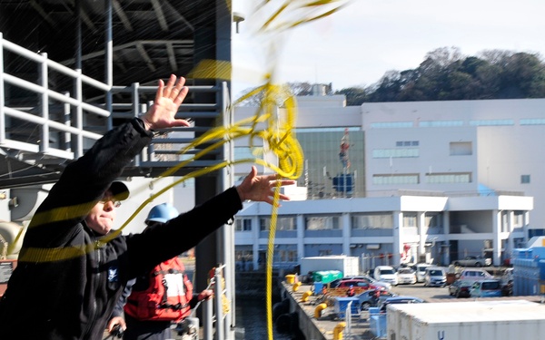 A sailor dressed in black is throwing a yellow heaving line from the deck of a naval vessel toward a pier or dock. A second crew member in a red jacket stands nearby observing. The scene appears to take place at a naval installation, likely in Japan based on the visible 'UNIX' container markings and architectural style.