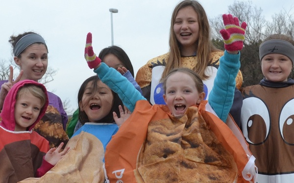 A group of children and at least one adult are dressed in food-themed costumes, appearing to participate in a fun run or themed event outdoors. The costumes depict peanut butter-related food items, including what appears to be a peanut butter sandwich, a peanut butter sardine, and a chocolate peanut butter cup character. The group is energetic and celebratory, posing with raised hands and big smiles.