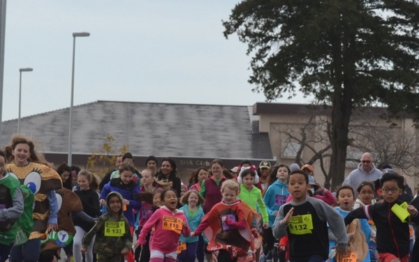 A large group of children and some adults are participating in a fun run or costume race on a paved road. Participants are wearing colorful costumes and yellow race bibs with visible numbers. The event appears to take place in a suburban or institutional setting, likely during autumn based on the bare trees and overcast sky.
