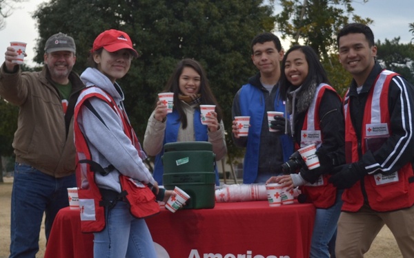 A group of six volunteers are gathered around an American Red Cross table outdoors, holding up cups in a celebratory pose. Several volunteers are wearing red American Red Cross vests, and the table is covered with a red American Red Cross tablecloth. The setting appears to be a park or open outdoor area during cooler weather.