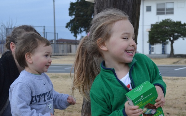 A young girl in a green fleece jacket is smiling and holding a box of Girl Scout cookies outdoors. She is accompanied by a toddler in a Pittsburgh sweatshirt and an adult woman crouching behind them. The setting appears to be a grassy area near institutional or military-style buildings.